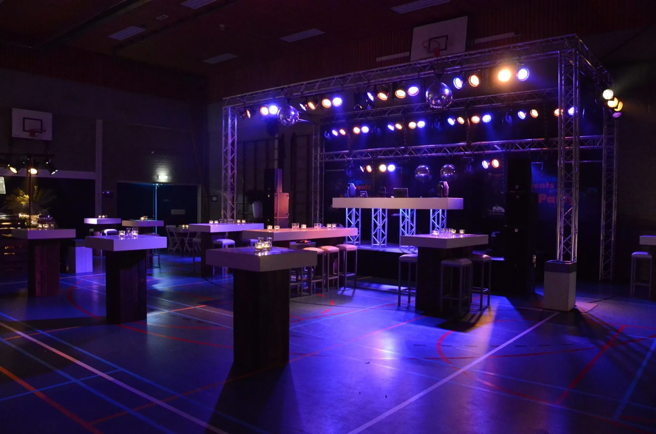 Sporthal omgebouwd tot feestzaal met statafels, DJ-booth en truss vol podiumlicht